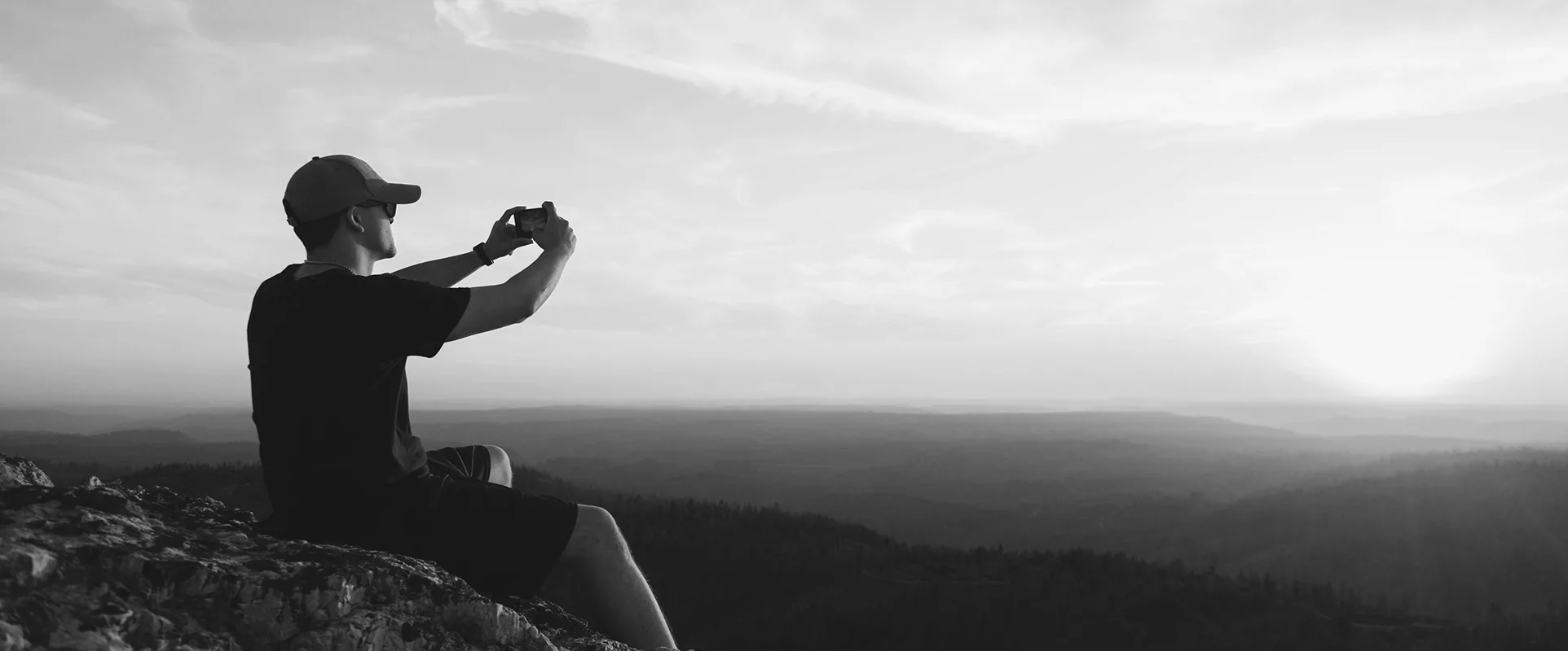 man taking a selfie or a photo, sitting at a ledge overlooking a sunset